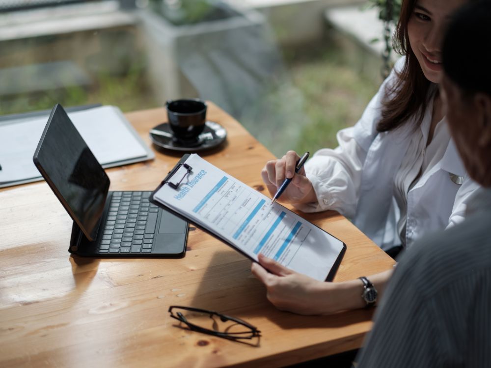 Plano de saúde é pago antecipadamente: Consultora explicando opções de planos de saúde para cliente, com formulário de seguro saúde em uma prancheta.